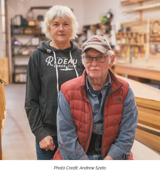 Joan Barrett and Ted Moores pose for a portrait against the backdrop of the Bear Mountain Boats workshop. A caption underneath "Photo Credit: Andrew Szeto"